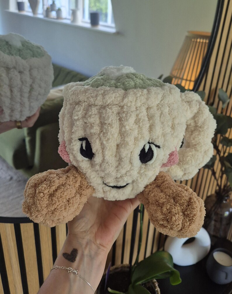 Pu&ograve; includere: Una tazza in crochet di peluche color crema con una parte superiore verde chiaro, due braccia in crochet marroni e guance rosa. La tazza presenta occhi neri ricamati e una piccola bocca nera, creando un volto amichevole. La texture &egrave; morbida.