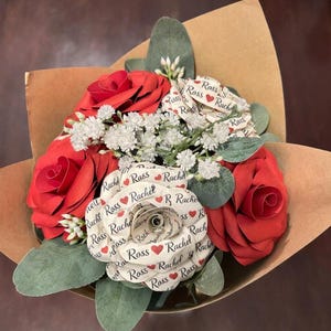 May include: A bouquet of paper flowers wrapped in brown paper. The bouquet features red paper roses and white paper flowers with the names "Rachel" and "Ross" printed on them. Green leaves and white filler flowers are also included.