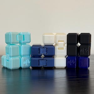 May include: A collection of colorful fidget cubes in shades of light blue, navy blue, cream, and black. Each cube is composed of smaller, connected blocks, creating a modular design. The cubes are arranged on a dark surface.