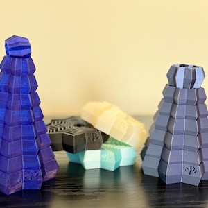 Puede incluir: Una colección de objetos geométricos impresos en 3D en varios colores. Los objetos incluyen dos estructuras altas en forma de cono en azul y gris, y varias piezas más pequeñas en forma de estrella en negro, blanco y turquesa. Los objetos están sobre una superficie oscura.