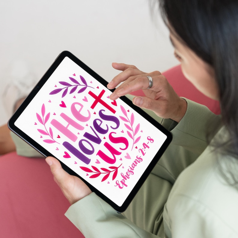 May include: A digital tablet screen displaying a graphic design with the text He loves us and Ephesians 2:4-8 in pink, purple, and red. The design features stylized leaf branches and small heart shapes surrounding the lettering on a white background.