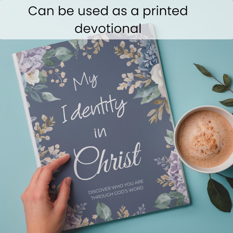 May include: A rectangular book with a dark blue cover featuring a floral border in shades of purple, white, and green. The text reads My Identity in Christ and Discover who you are through God's word. A white mug with a light brown beverage sits nearby.