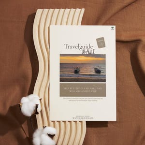 Può includere: Una guida turistica intitolata "Travelguide BALI" con una foto del tramonto di barche sulla copertina. Il libro è sostenuto da un elemento decorativo in legno chiaro e steli di cotone. Il testo recita "Step by Step to a Relaxed and Well-Organized Trip."