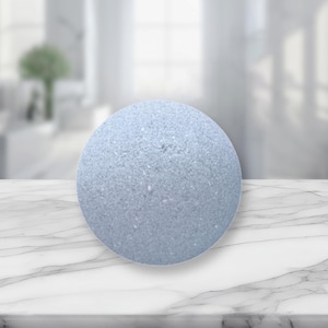 以下が含まれることがあります： テクスチャ加工された表面を持つ、ライトブルーの丸いバスボム。バスボムは白い大理石の表面に置かれています。