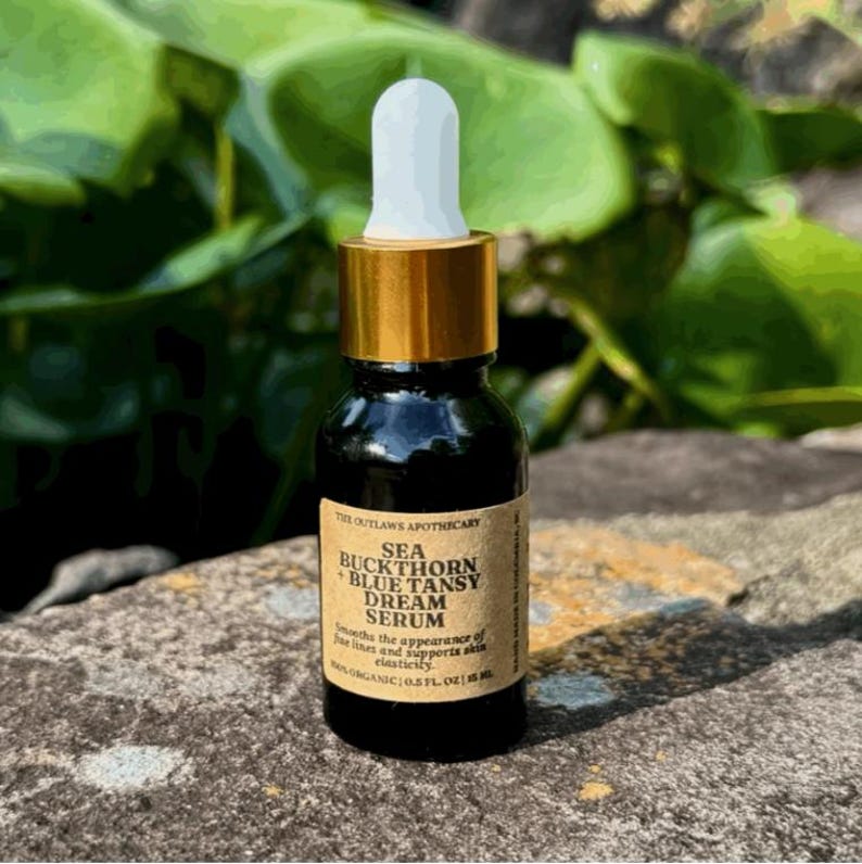 May include: A small, dark glass bottle of Sea Buckthorn Blue Tansy Dream Serum with a white dropper and gold cap. The label reads "The Outlaws Apothecary" and "Organic 0.5 fl oz". The bottle is on a stone surface with green foliage in the background.
