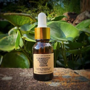May include: A brown glass bottle with a gold cap and white dropper, labeled "FERMENTED ROSEHIP+ NEROLI GLOW SERUM." The bottle is 0.5 fl oz. The background features green foliage and a stone surface.