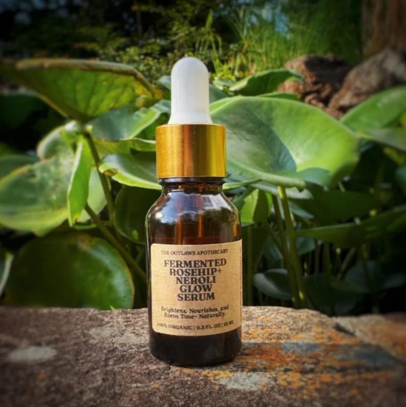 May include: A brown glass bottle with a gold cap and white dropper, labeled "FERMENTED ROSEHIP+ NEROLI GLOW SERUM." The bottle is 0.5 fl oz. The background features green foliage and a stone surface.