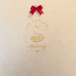 May include: A light beige paper bag with a gold logo that reads "THE OUTLAWS Apothecary" and a red satin bow at the top. The logo features a crescent moon and a snake.