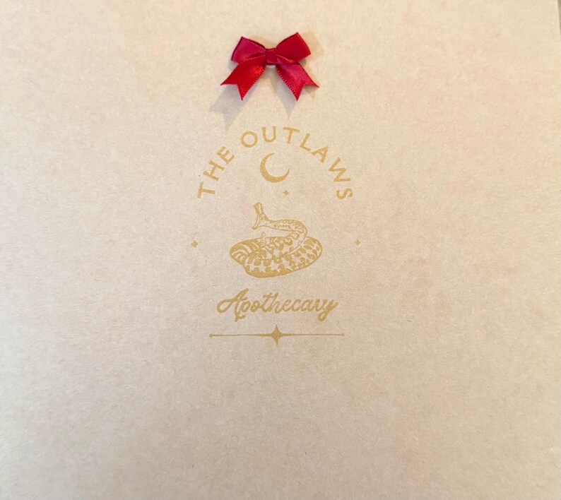 May include: A light beige paper bag with a gold logo that reads "THE OUTLAWS Apothecary" and a red satin bow at the top. The logo features a crescent moon and a snake.