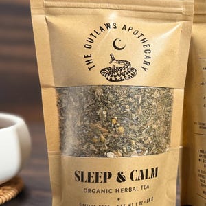 May include: A brown paper bag of "Sleep & Calm" organic herbal tea. The bag features the text "The Outlaws Apothecary" with a snake and crescent moon logo. The bag contains a mix of dried herbs and is labeled "Caffeine-Free".