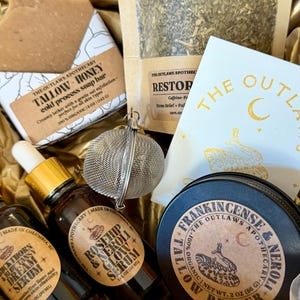 May include: A gift box filled with apothecary products. Items include a soap bar, a bag of herbs, a serum bottle, a tin of tallow butter, and a tea infuser. The products have labels with text such as "Tallow Honey" and "Rosehip & Neroli Glow Serum".
