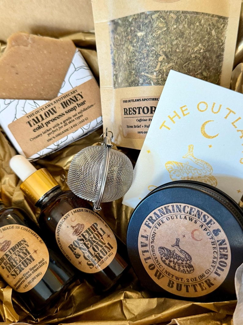 May include: A gift box filled with apothecary products. Items include a soap bar, a bag of herbs, a serum bottle, a tin of tallow butter, and a tea infuser. The products have labels with text such as "Tallow Honey" and "Rosehip & Neroli Glow Serum".