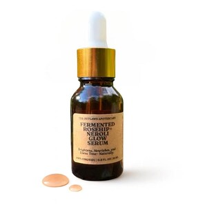 May include: A brown glass bottle of The Outlaws Apothecary Fermented Rosehip + Neroli Glow Serum. The bottle has a gold cap and a white dropper. The label reads "Brightens, Nourishes, and Evens Tone- Naturally". Two small drops of serum are next to the bottle. The bottle contains 0.5 fl oz.