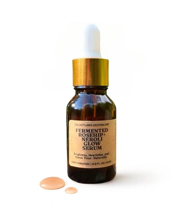May include: A brown glass bottle of The Outlaws Apothecary Fermented Rosehip + Neroli Glow Serum. The bottle has a gold cap and a white dropper. The label reads "Brightens, Nourishes, and Evens Tone- Naturally". Two small drops of serum are next to the bottle. The bottle contains 0.5 fl oz.