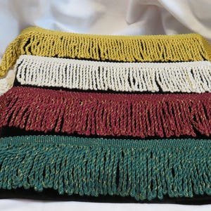 May include: Four strips of decorative fringe trim layered on a black surface. The fringe colors from top to bottom are metallic gold, cream, deep red with gold threading, and dark green with gold threading. Each strip features twisted cord tassels.