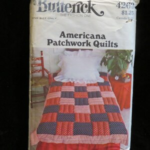 May include: A vintage sewing pattern titled "Americana Patchwork Quilts" by Butterick. The pattern cover shows a bed with a patchwork quilt in red, white, and blue. A white pillow with a ruffled edge rests on the bed.