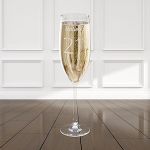 Puede incluir: Una copa de champán llena de un líquido dorado y burbujeante. La copa está grabada con el nombre "Hailey's" y el número "21". La cristalería elegante es ideal para una celebración.