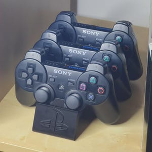 Puede incluir: Una colección de mandos negros Sony PlayStation 3 DualShock 3, apilados y expuestos en una estantería de madera. Cada mando presenta el logotipo de Sony, las palabras select, start y DualShock 3. Los mandos tienen botones de acción de colores y un soporte negro con el logotipo de PlayStation.