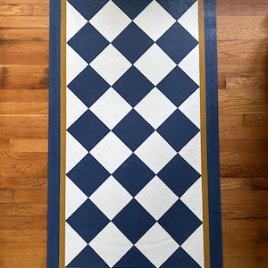 May include: Rectangular rug with a navy blue border and a gold inner border. The rug's central design features a pattern of white and navy blue diamonds. The rug is placed on a hardwood floor.