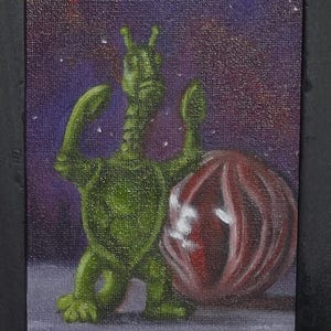 Op de afbeelding: Een ingelijst schilderij toont een groene alienfiguur met opgeheven armen naast een rood-wit gestreepte bol. De achtergrond is een sterrenhemel van paars en blauw. Het frame is zwart.