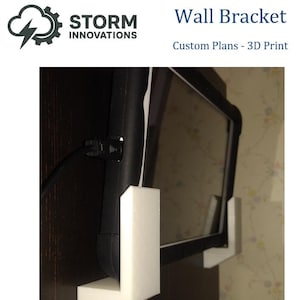 May include: A white 3D-printed wall bracket holding a black tablet. The bracket is mounted on a dark brown surface. The text "Wall Bracket" and "Custom Plans - 3D Print" are visible.