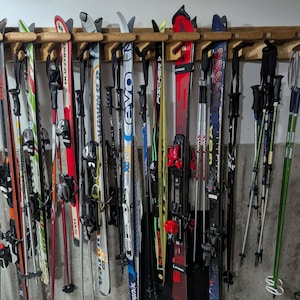 Puede incluir: Un estante de esquí de madera que contiene una variedad de esquís y bastones de esquí. Los esquís son de diferentes colores y diseños, incluyendo rojo, verde y azul. Los bastones de esquí son negros y verdes. El estante está montado en una pared de hormigón gris.