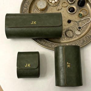 May include: Three olive green leather cases with gold lettering "JK". The cases are in different shapes and sizes, including a rectangular case, a square case, and a cylindrical case. The cases are on a silver tray with jewelry.