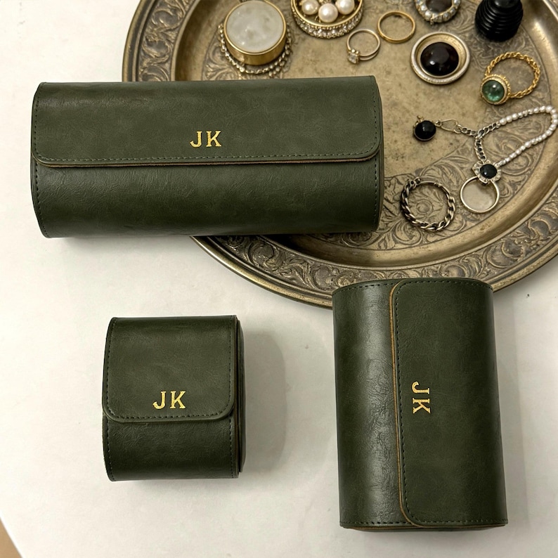 May include: Three olive green leather cases with gold lettering "JK". The cases are in different shapes and sizes, including a rectangular case, a square case, and a cylindrical case. The cases are on a silver tray with jewelry.