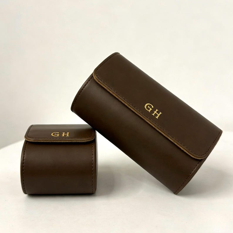 May include: Two brown leather watch cases, one cylindrical and one square, with gold-colored initials "GH" embossed on the front. The cylindrical case is leaning against the square case.