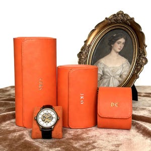 May include: An orange leather watch case set with a mechanical watch and three additional cases. The watch has a black band and a silver face. The cases have gold lettering: LERS, JKO, and DC.
