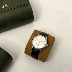 May include: A silver wristwatch with a white face and black leather band rests on a brown cushion. The watch face features Roman numerals and the initials "JK". A dark green leather case with the initials "JK" is in the background.