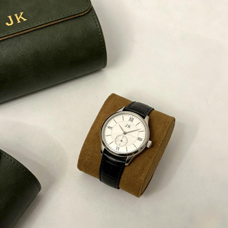 May include: A silver wristwatch with a white face and black leather band rests on a brown cushion. The watch face features Roman numerals and the initials "JK". A dark green leather case with the initials "JK" is in the background.