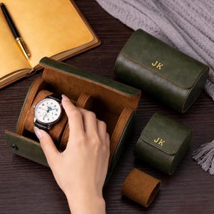 May include: A green leather watch case with a brown interior, holding a silver watch with a black band. Two additional green cases and a watch roll are also present. The initials "JK" are embossed in gold on the cases.