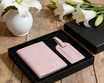 May include: A blush pink passport holder and luggage tag set, presented in a black gift box. Both items are embossed with the initials "TU". White lilies in a white vase and a black box are in the background.
