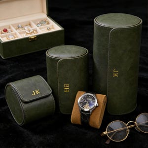 May include: An assortment of green leather watch cases and a jewelry box. The watch cases are cylindrical and rectangular, with gold initials. A wristwatch with a black leather band sits on a brown stand. Gold-rimmed glasses are also visible.
