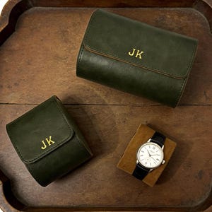 Può includere: Custodie per orologi in pelle verde scuro con iniziali dorate "JK" e un orologio da polso con cinturino in pelle nera e cassa argentata. L'orologio è esposto su un cuscino di velluto marrone, il tutto su un vassoio di legno.