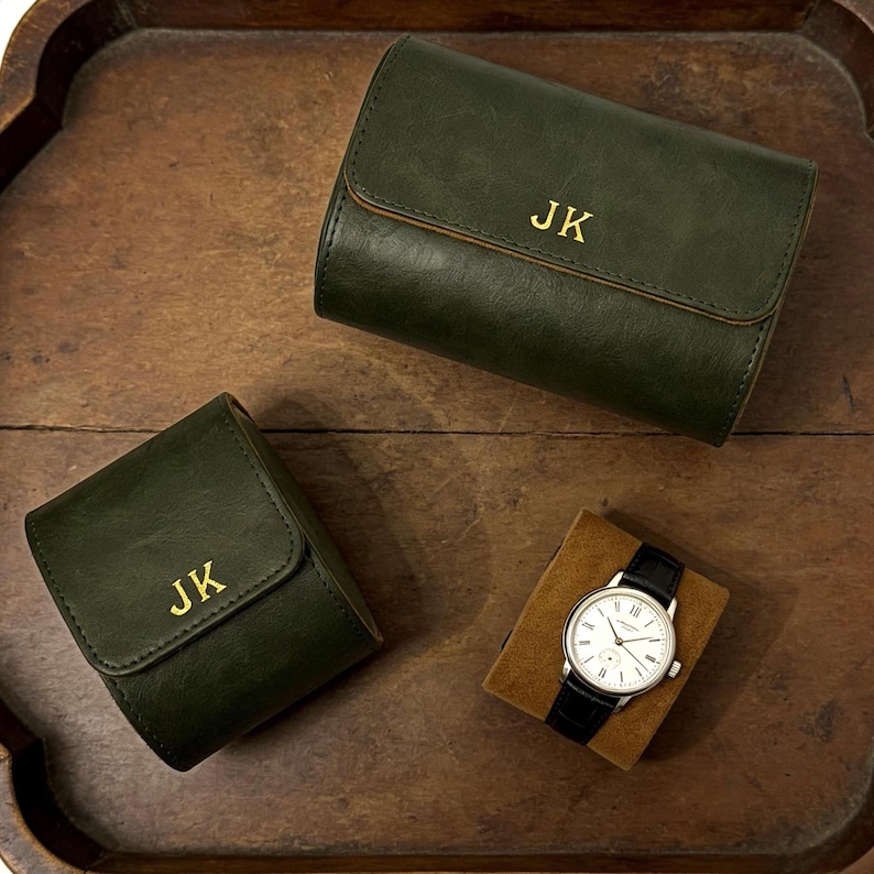 May include: Dark green leather watch cases with gold initials "JK" and a wristwatch with a black leather band and silver case. The watch is displayed on a brown velvet cushion, all on a wooden tray.