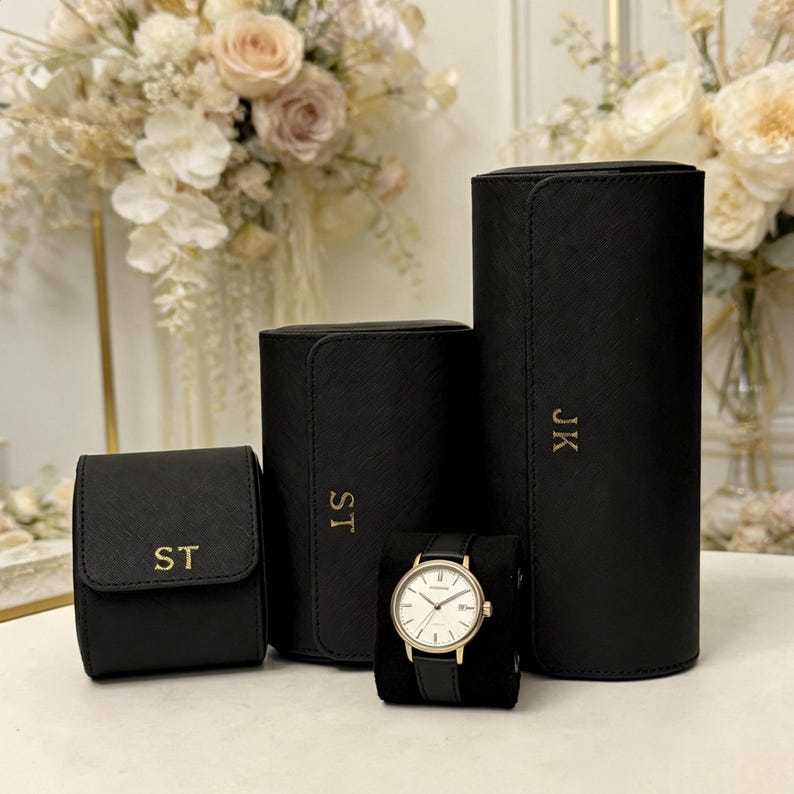 May include: A collection of black watch cases with gold initials, alongside a gold-toned watch with a black leather band. The cases vary in size and shape, with a small square case, a medium rectangular case, and a tall cylindrical case.