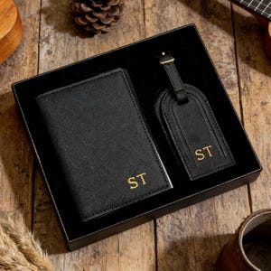 以下が含まれることがあります： 金色のイニシャル「ST」が入った黒いパスポートケースとラゲッジタグのギフトセット。旅行用アクセサリーに最適な黒い箱に入っています。木製の表面にセットが置かれています。