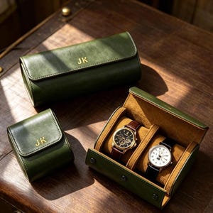 May include: Three olive green watch cases with gold-colored JK monograms. One long case is closed, one small square case is closed, and one rectangular case is open, displaying two wristwatches with leather straps on tan cushions.