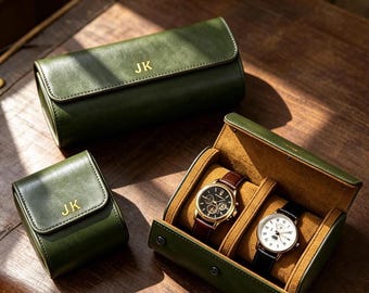Könnte beinhalten: Drei olivgrüne Uhrenetuis mit goldenen JK-Monogrammen. Ein langes Etui ist geschlossen, ein kleines quadratisches Etui ist geschlossen und ein rechteckiges Etui ist geöffnet und zeigt zwei Armbanduhren mit Lederarmbändern auf beigen Kissen.
