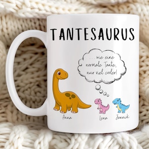 May include: White ceramic mug with the word "TANTESAURUS" in black. The mug features a cartoon dinosaur and three smaller dinosaurs in pink and blue. Text in a thought bubble reads "...wie eine normale Tante, nur viel cooler!" with names below the dinosaurs.