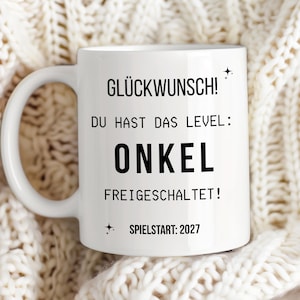 Könnte beinhalten: Eine weiße Keramiktasse mit schwarzem Aufdruck: GLÜCKWUNSCH! DU HAST DAS LEVEL: ONKEL FREIGESCHALTET! SPIELSTART: 2027, ergänzt durch kleine Sternsymbole. Die Tasse hat einen geschwungenen Henkel auf der linken Seite und eine glatte Oberfläche.