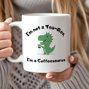 Könnte beinhalten: Weiße Keramik-Tasse mit dem Text "I'm not a Tea-Rex, I'm a Coffeesaurus" und einer grünen Dinosaurier-Illustration, die eine Kaffeetasse hält. Die Tasse hat einen weißen Griff.