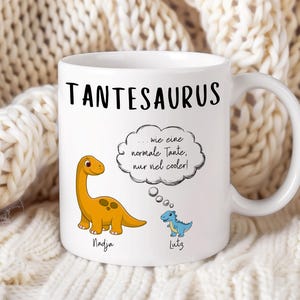 May include: White ceramic mug with the word "TANTESAURUS" in black. Features a cartoon orange dinosaur and a blue dinosaur, with text in a thought bubble. The names "Nadja" and "Lutz" are below the dinosaurs.