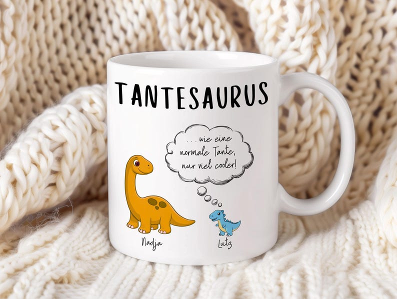 May include: White ceramic mug with the word "TANTESAURUS" in black. Features a cartoon orange dinosaur and a blue dinosaur, with text in a thought bubble. The names "Nadja" and "Lutz" are below the dinosaurs.