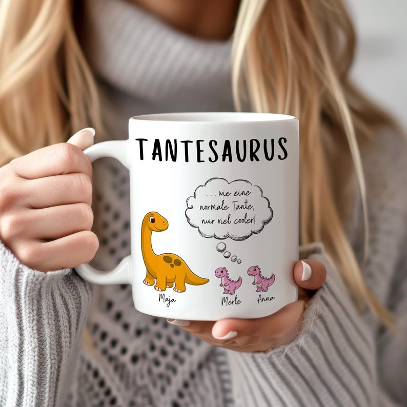 May include: White ceramic mug with the word "TANTESAURUS" in black. The mug features a cartoon orange dinosaur and two pink dinosaurs. Text in German reads "...wie eine normale Tante, nur viel cooler!" with the names Maya, Merle, and Anna.