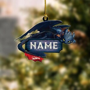May include: A black dragon ornament with a red tail fin and the word "NAME" on a wooden sign. The dragon is lying down, with yellow eyes and a harness. The ornament hangs from a gold string.