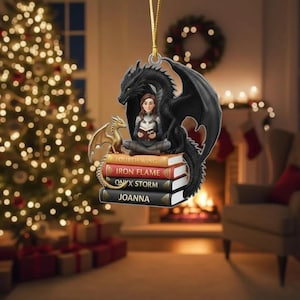 May include: A Christmas ornament featuring a woman reading atop a stack of books, with a black dragon and a smaller gold dragon. The books are titled "Fourth Wing", "Iron Flame", "Onyx Storm", and "Joanna". The ornament hangs from a gold string.