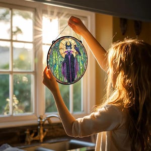 Puede incluir: Arte de ventana de vidriera redonda con un personaje morado y negro con un cuervo. La obra de arte tiene un borde negro y est&aacute; suspendida por una cuerda negra. El fondo incluye un dise&ntilde;o de vidriera con tonos verdes y amarillos.